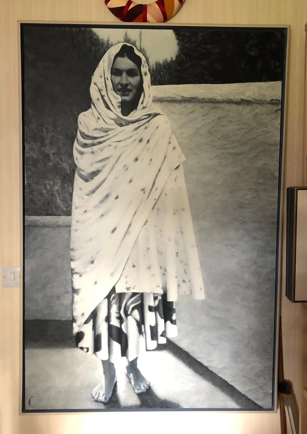A large black and white painting of a woman standing barefoot on a terrace, wrapped in a pale spotted shawl over a patterned skirt, with a long shadow across the paving.