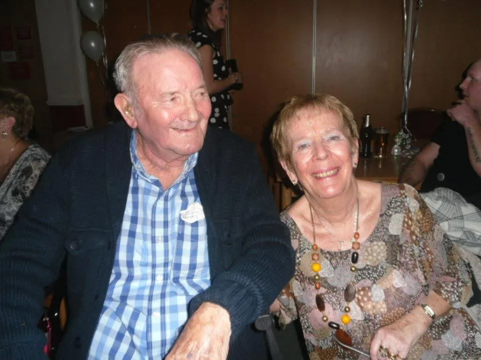 An older man in a blue checked shirt and cardigan sitting beside an older woman in a patterned top and beaded necklace, both smiling, with balloons behind them