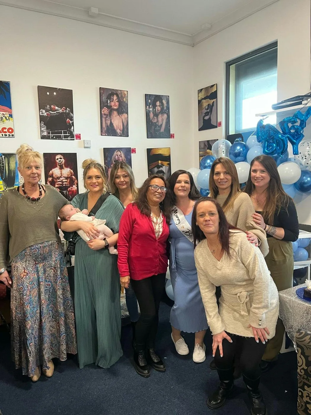 A group of women posing together in a room decorated with blue and white balloons, one of them holding a baby